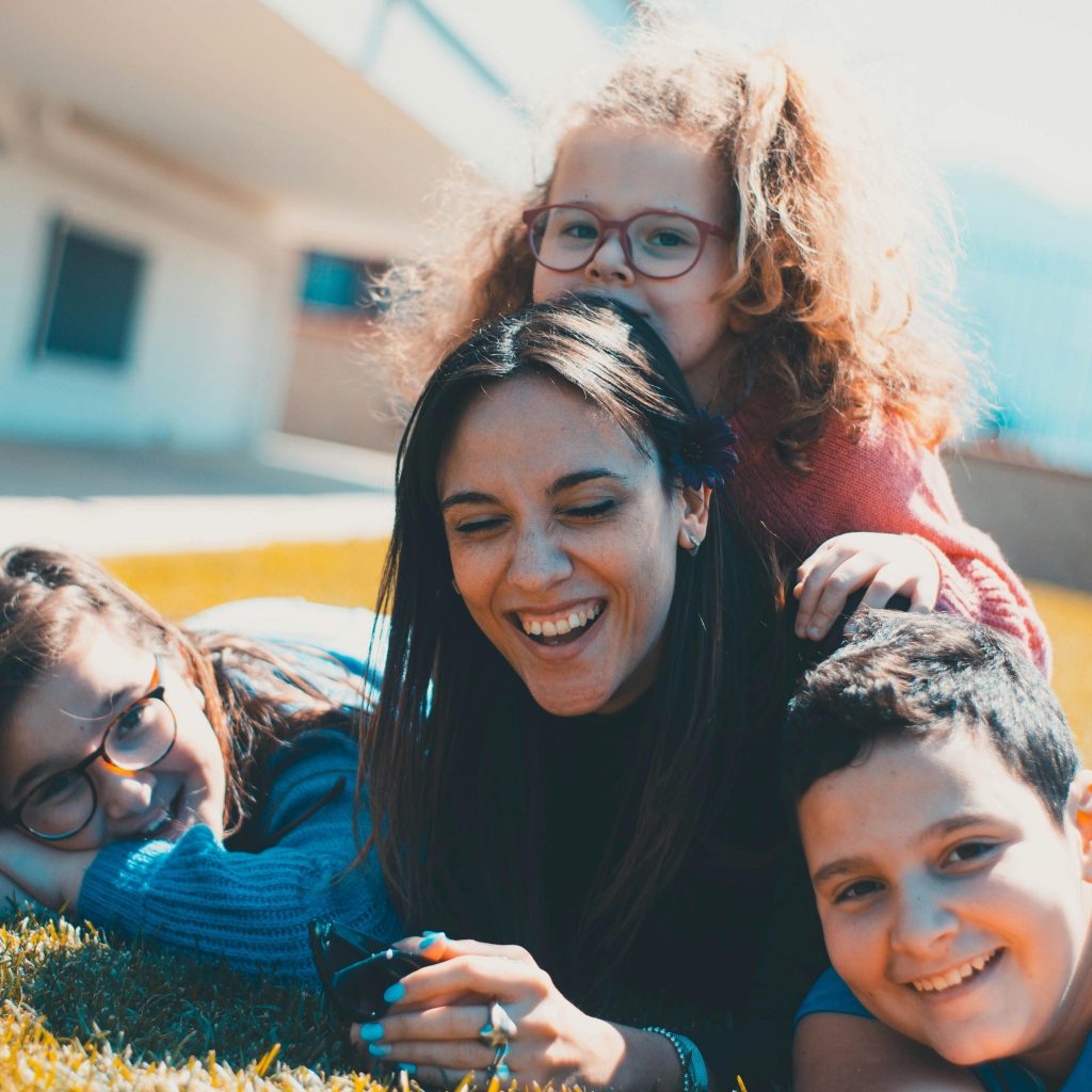 Mother and children laughing together on the grass, enjoying a sunny day outdoors.
